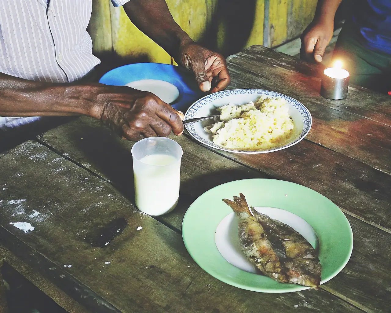 Weathered hands sharing a simple meal by candlelight in a dim Amazon village